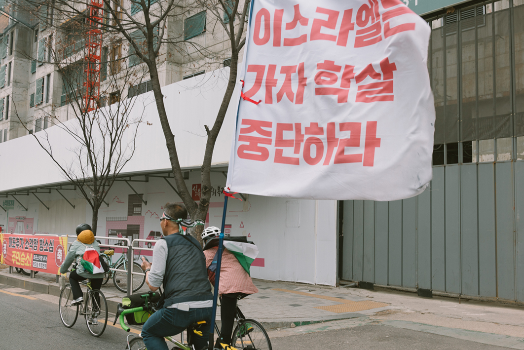 세 대의 자전거가 도로를 달리는 모습을 촬영한 사진이다. 앞선 두 자전거 탑승자는 몸에 팔레스타인 국기를 부착하고 있고, 뒤쪽 자전거 탑승자는 머리에 팔레스타인 국기 문양의 머리띠를 두르고 '이스라엘은 가자 학살 중단하라'라는 문구가 적힌 큰 깃발을 자전거에 달고 있다.