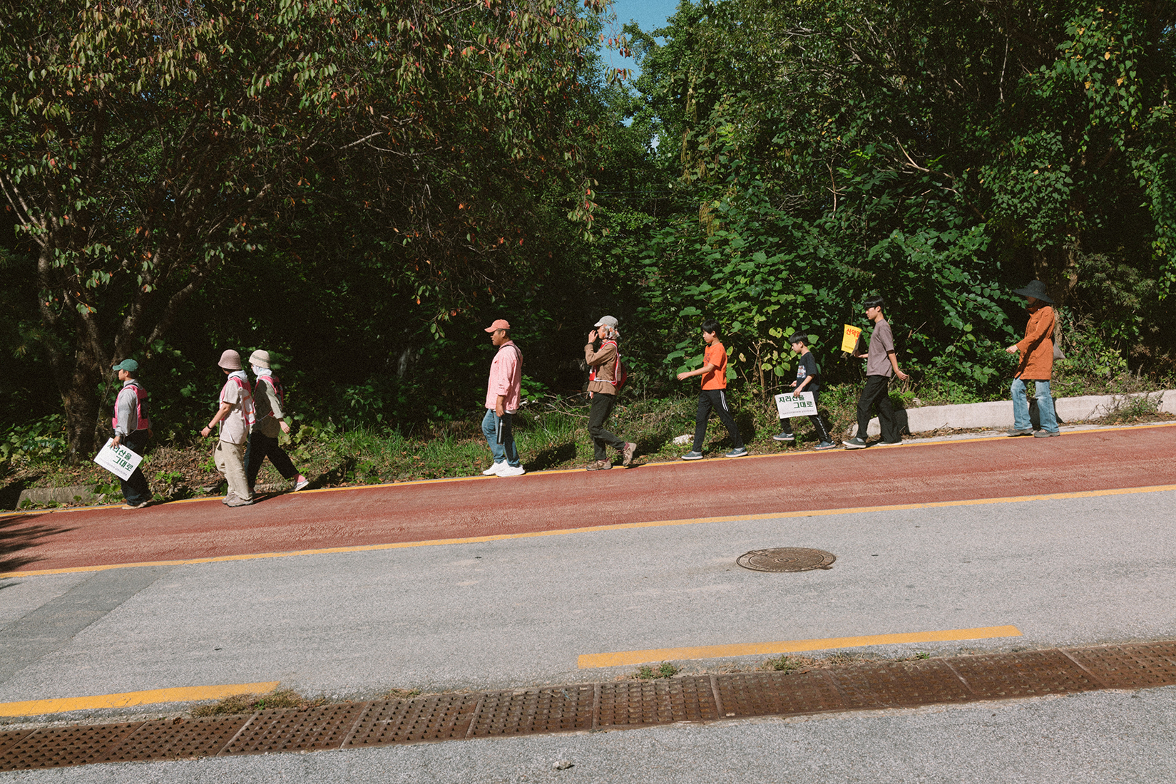 내리막 도로 옆으로 몇몇 사람들이 걸어 내려오고 있다. 사람들 손과 몸에는 ‘지리산 그대로’, ‘산악열차 백지화’ 등의 문구가 적힌 피켓과 배너가 있다. 도로 옆으로는 풀과 나무가 우거져있다. 
