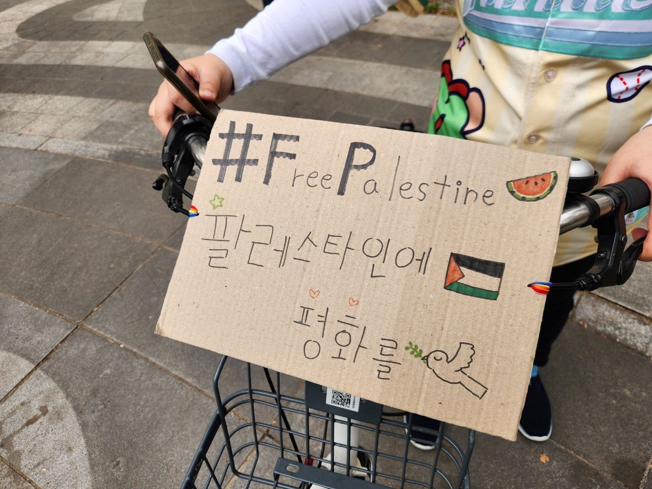 자전거 핸들 앞에 골판지로 만든 피켓이 부착된 모습을 촬영한 사진이다. 피켓에는 '#Free Palestine. 팔레스타인에 평화를'이라는 문구와 수박, 팔레스타인 국기, 올리브가지를 문 비둘기 등의 그림이 그려져있다.
