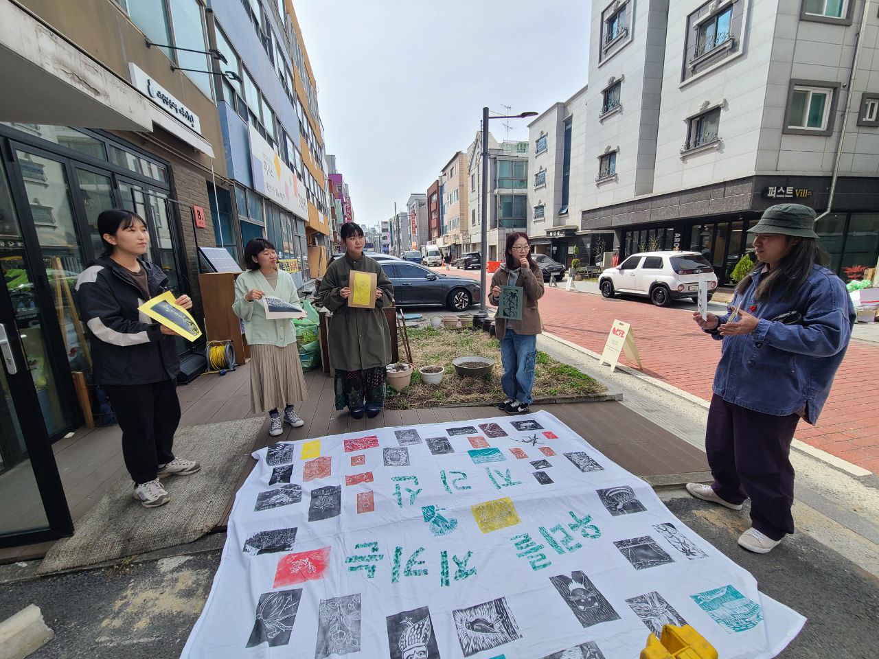 길 한켠 바닥에 '우리를 지키는 지리산'이라는 문구와 여러색의 판화가 찍힌 큰 천이 놓여있고 그 주변에 4명의 사람들이 종이에 찍힌 작은 판화를 들고 서 있다. 