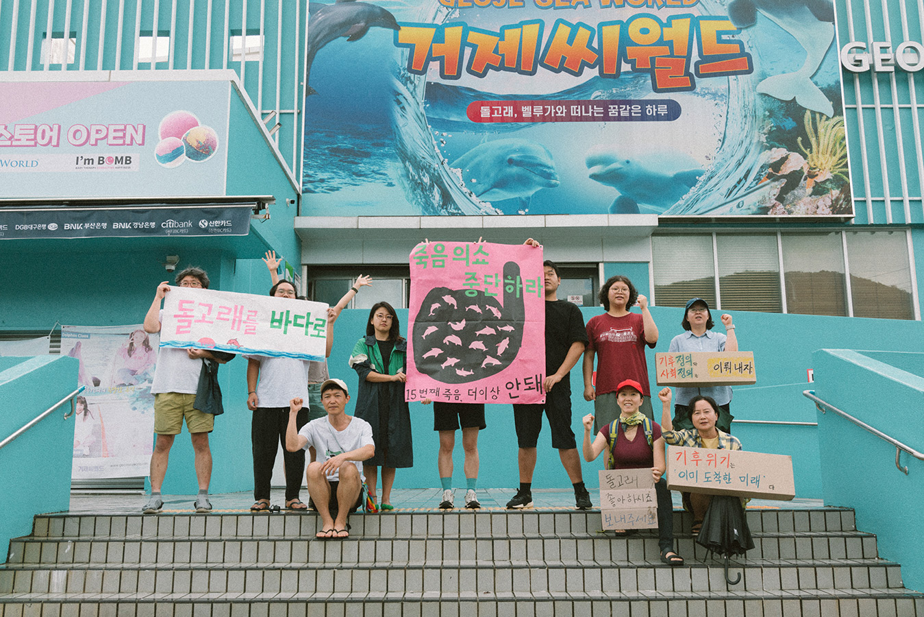 거제씨월드 앞 계단에서 10명의 사람들이 "돌고래를 바다로, 죽음의 쇼 중단하라, 15번째 죽음 더이상 안돼, 기후위기 이미 도착한 미래, 돌고래 좋아하시죠 보내주세요' 등의 문구가 적힌 피켓과 배너를 들고 서거나 앉아서 주먹을 들거나 팔을 뻗고 있다