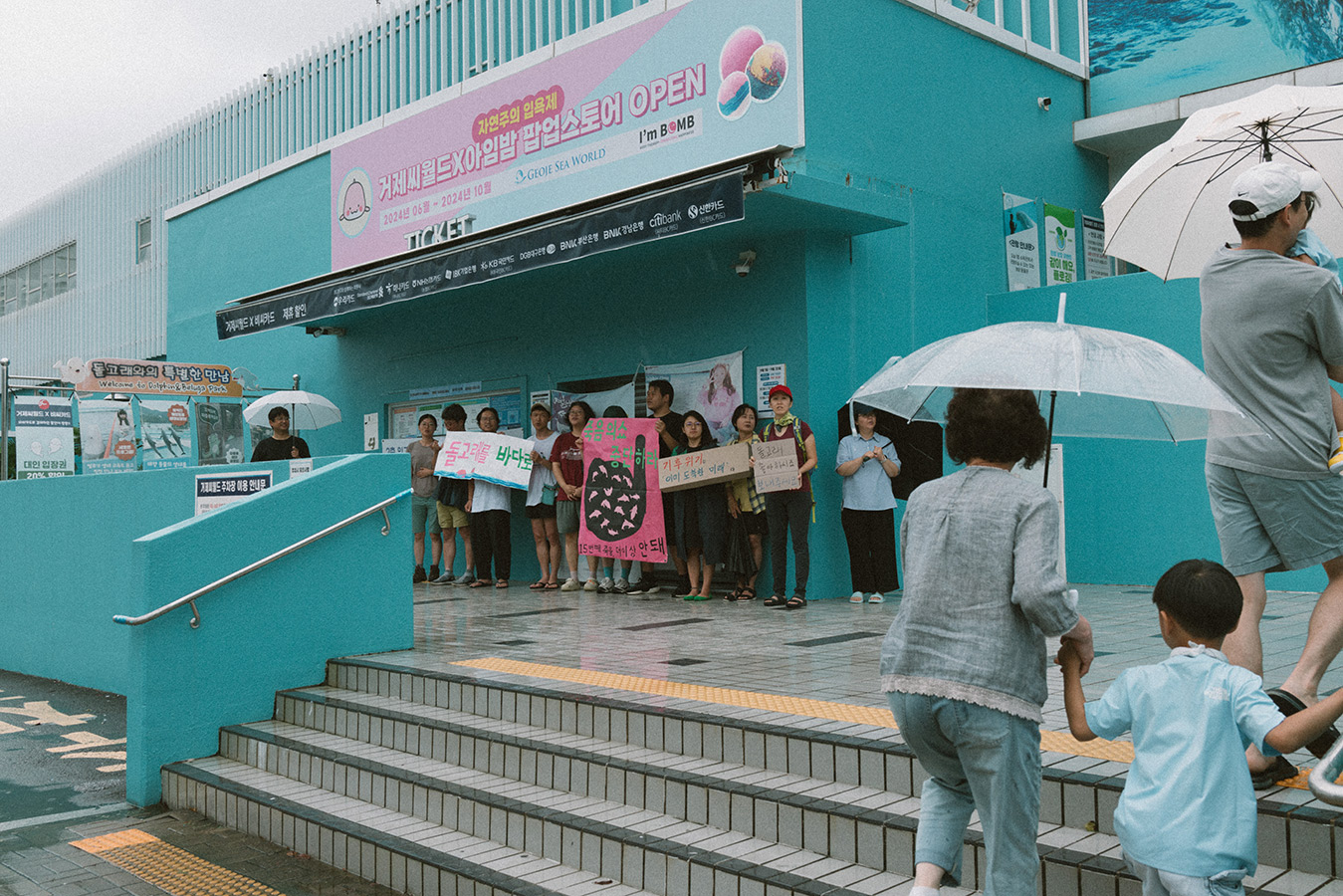 시설물 계단 벽면 쪽에 10여명의 사람들이 "돌고래를 바다로, 죽음의 쇼 중단하라, 15번째 죽음 더이상 안돼, 기후위기 이미 도착한 미래, 돌고래 좋아하시죠 보내주세요' 등의 문구가 적힌 피켓과 배너를 들고 서 있다.  계단 쪽에서 우산을 쓴 가족들이 피켓과 배너를 든 사람들을 쳐다보며 지나간다.