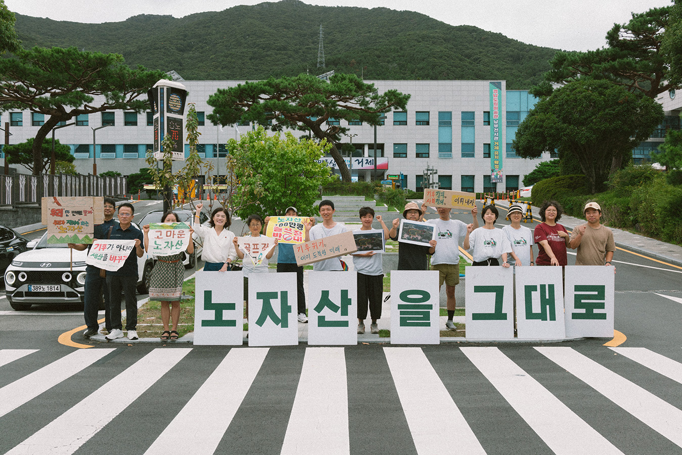 관공서 진입로 부근 공터에서 14명의 사람들이 '노자산을 그대로, 고마운 노자산, 기후가 아니라 세상을 바꾸자, 기후위기 이미 도착한 미래, 기후정의 사회정의 이뤄내자, 우리만 편해도 될까 우리가 지켜야 하는 노자산' 등의 문구가 적힌 다양한 현수막과 피켓을 들고 서 있다.