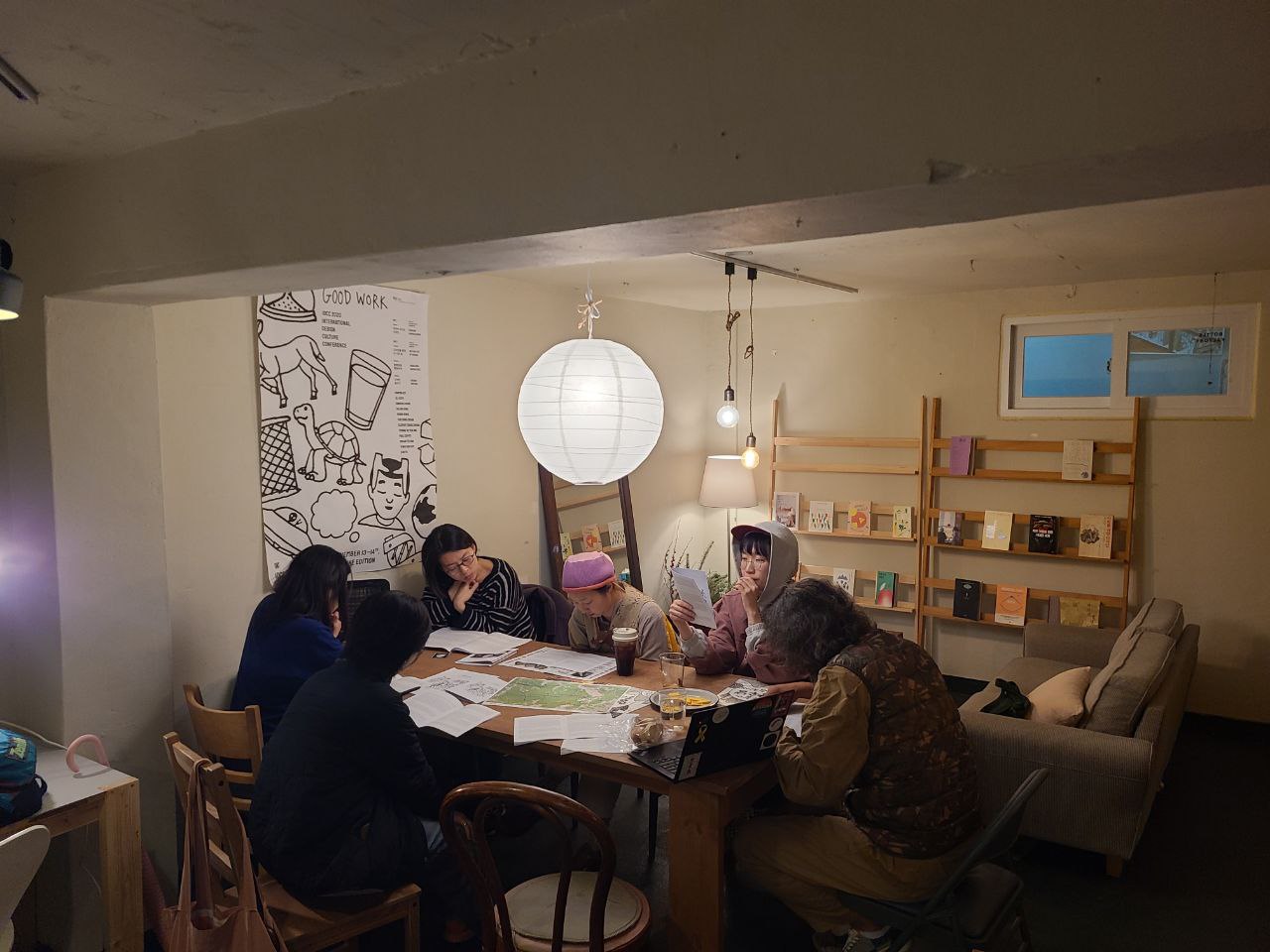  책장과 소파가 있는 실내에 6명의 사람들이 테이블에 둘러앉아 소책자를 보고 있다. 테이블 위에는 지도와 자료들이 놓여있다. 