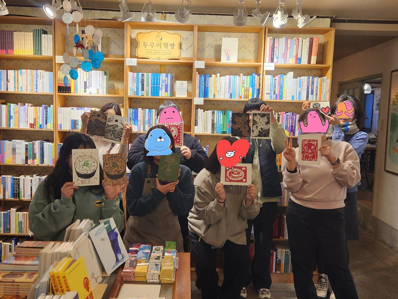 책방 실내에서 8명의 사람들이 작은 판화를 들고 촬영한 사진이다. 뒤쪽 책장에는 많은 책이 꽂혀있고, 위쪽에 '두루미 책방'이라는 간판이 놓여있다. 사람들은 각자 목판화 또는 목판화와 목판을 들고 서 있으며 사람들의 얼굴은 판화나 스티커로 가려져있다. 