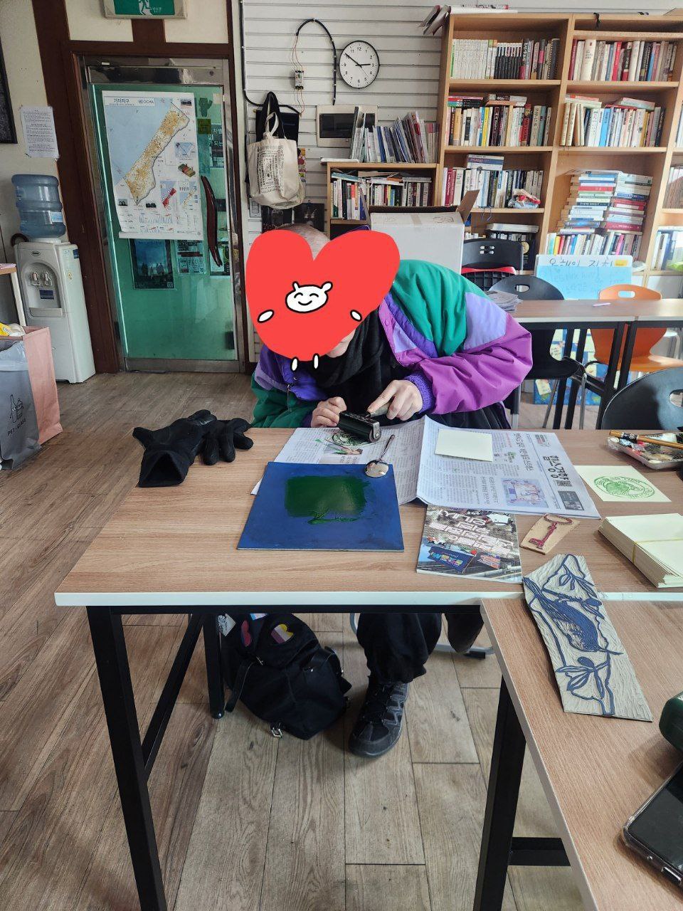 1명의 사람이 실내의 테이블 위에서 판화를 찍고 있는 모습을 촬영한 사진이다. 얼굴은 스티커로 가려져있다. 