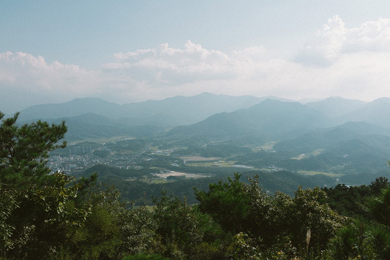 산 아래 시가지 풍경이 보이는 사진이다. 뒤로 높은 산들이 많고 하늘에는 구름이 조금 있다.